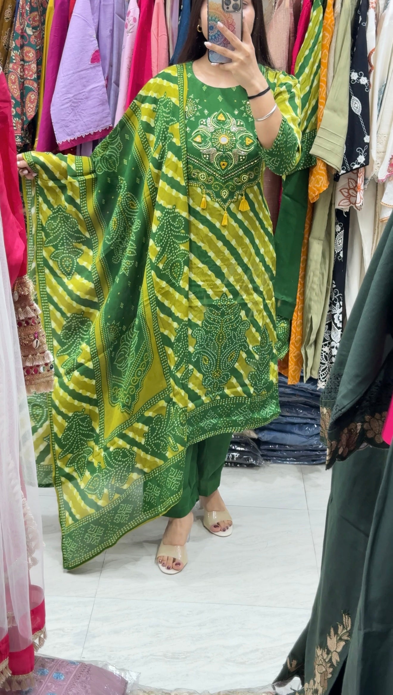A woman takes a mirror selfie in a clothing store while wearing a green and yellow tie-dye kurta with green pants. The background shows racks of various colorful garments.