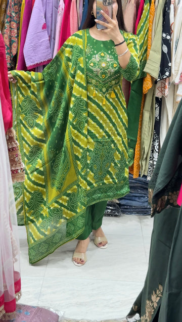 A woman takes a mirror selfie in a clothing store while wearing a green and yellow tie-dye kurta with green pants. The background shows racks of various colorful garments.
